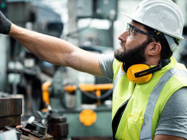 An industrial engineer wearing a white hard hat and yellow vest standing in an industrial factory, reaching for a machine.