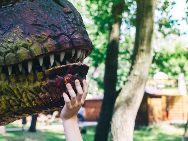 A child wearing a hat reaching out to touch the snout of an animatronic dinosaur in a fun theme park location.